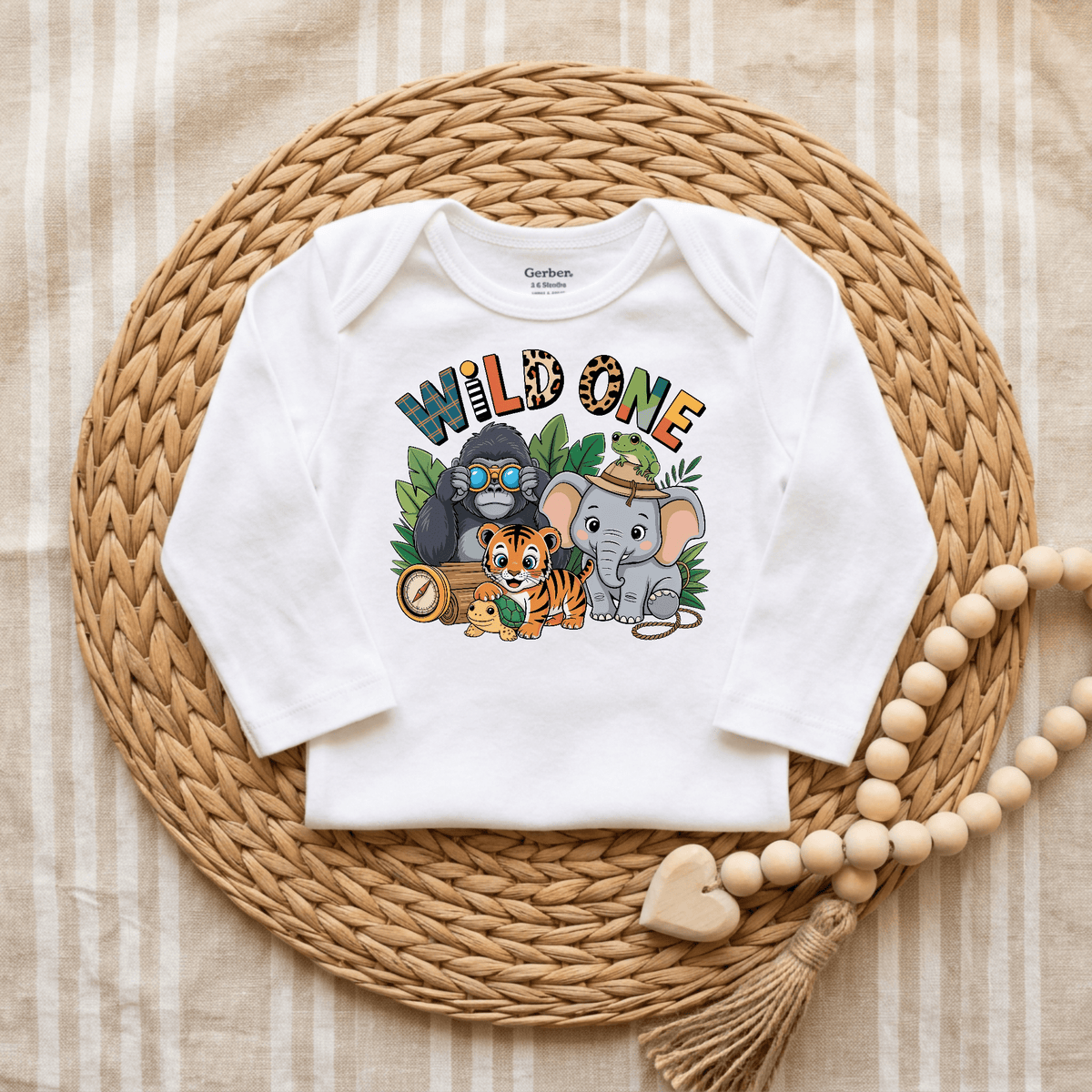Toodleroos Wild One 1st Birthday Baby Onesie – Jungle Safari Bodysuit in white, adorned with playful cartoon animals (gorilla, tiger, elephant) and leafy jungle accents. Ideal as a first birthday outfit for a safari-themed celebration. Shown displayed on a woven mat with a wooden bead garland alongside.