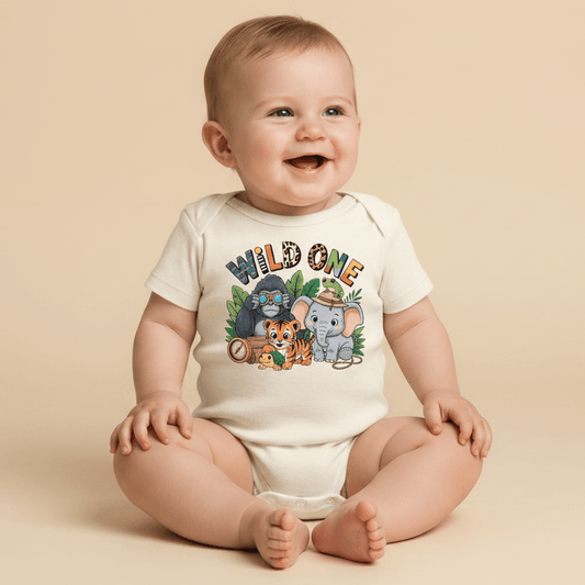 A happy baby sits on the floor wearing the Toodleroos Wild One 1st Birthday Baby Onesie – Jungle Safari Bodysuit, featuring cartoon animals like a lion, tiger, elephant, and koala. Surrounded by green leaves on a beige background, this adorable outfit is perfect for a safari-themed first birthday.