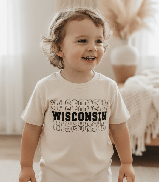 A smiling young child with light brown hair wears the Toodleroos Wisconsin Varsity Toddler Shirt – Kids State Pride Tee, standing in a cozy, softly lit room with neutral decor and a large vase in the background—a perfect scene capturing hometown pride.