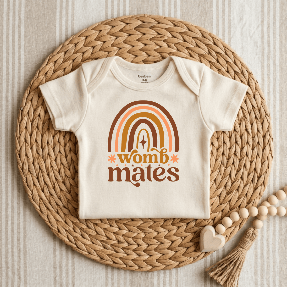 The Womb Mates Baby Onesie Set – Twin Boho Rainbow Bodysuits by Toodleroos features creamy baby bodysuits adorned with a retro-inspired rainbow and “womb mates” text in warm orange and brown hues, beautifully showcased on a woven mat beside a wooden bead toy—an adorable twin baby shower gift or an ideal nostalgic addition to any matching twin onesie collection.