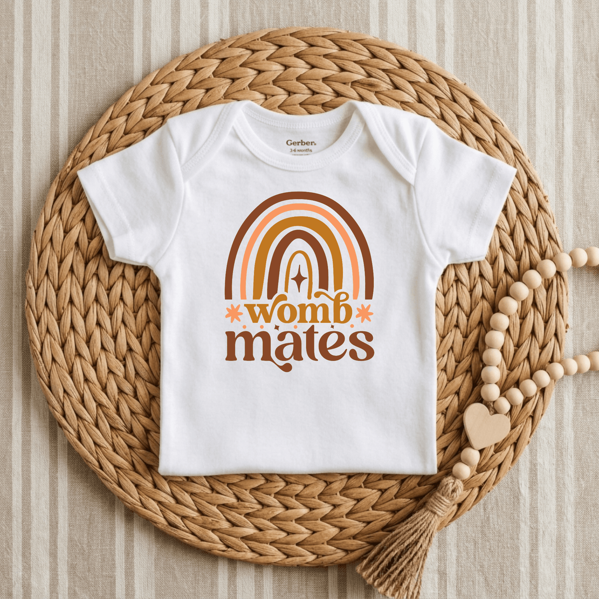 The Toodleroos Womb Mates Baby Onesie Set – Twin Boho Rainbow Bodysuits features white baby onesies adorned with a retro orange rainbow and “womb mates” printed on the front, styled on a woven mat with a beaded garland and striped fabric background—making them the perfect charming twin baby shower gift or addition to an adorable twin onesie set.