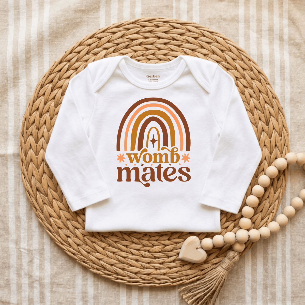 The Toodleroos Womb Mates Baby Onesie Set – Twin Boho Rainbow Bodysuits features a white bodysuit adorned with a retro rainbow and the phrase “womb mates,” displayed on a woven mat beside a beaded garland with a wooden heart, all set against a beige striped fabric backdrop—an ideal choice for an adorable twin onesie set or as a thoughtful twin baby shower gift.
