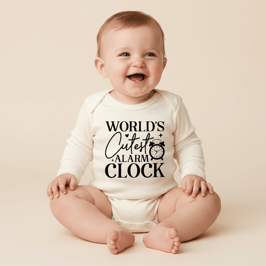 A smiling baby sits on the floor wearing the Toodleroos World’s Cutest Alarm Clock Onesie – a cream-colored, unisex baby bodysuit featuring the playful text “WORLDS CUTEST ALARM CLOCK” and a cute alarm clock illustration on the front—making it a hilarious baby shower gift parents are sure to adore.