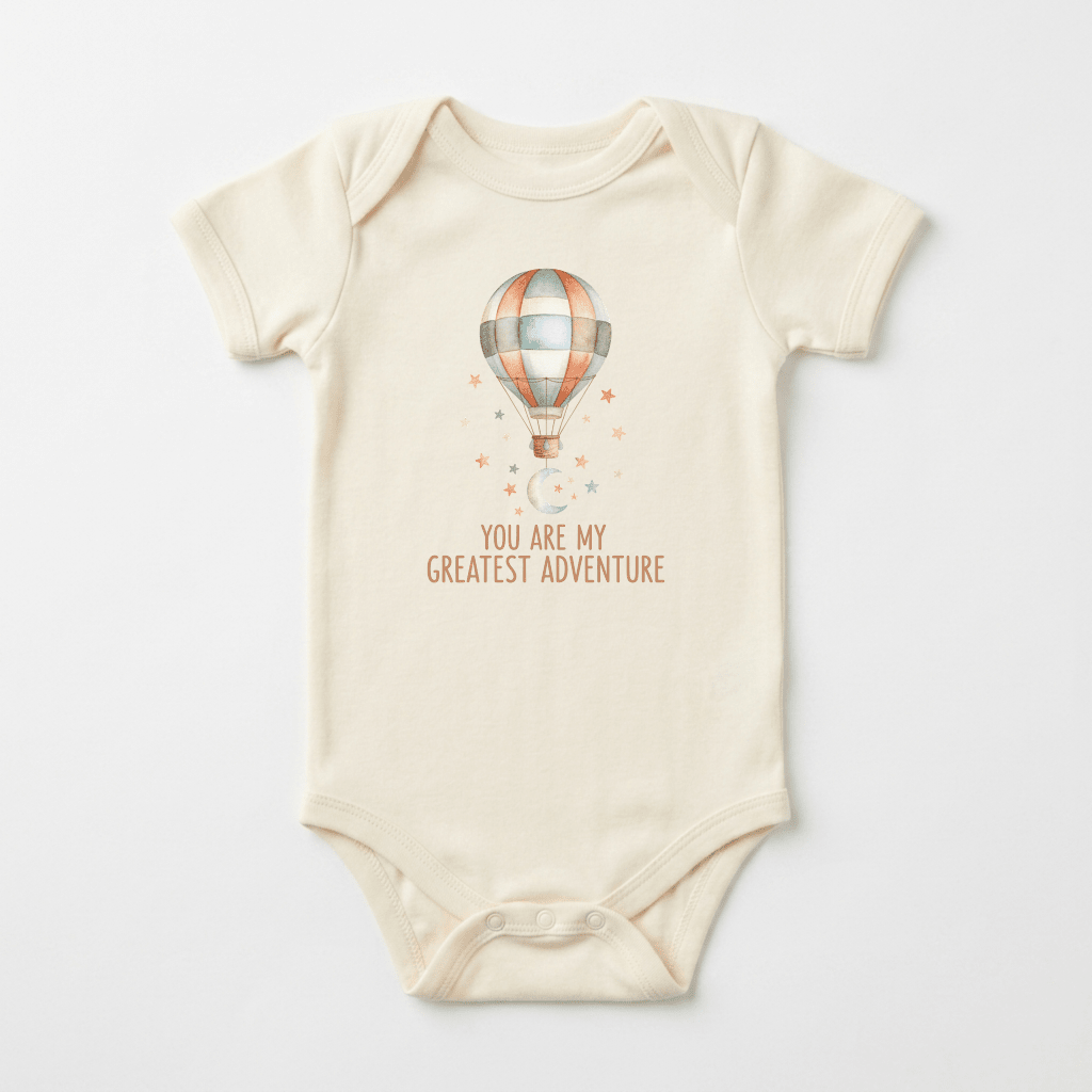 The Toodleroos “You Are My Greatest Adventure Baby Onesie – Hot Air Balloon” features a cream-colored bodysuit adorned with a charming hot air balloon above the phrase “You are my greatest adventure,” surrounded by tiny stars and moons. Laid flat on a white background, this adorable adventure-themed onesie makes a perfect gift for welcoming little explorers.
