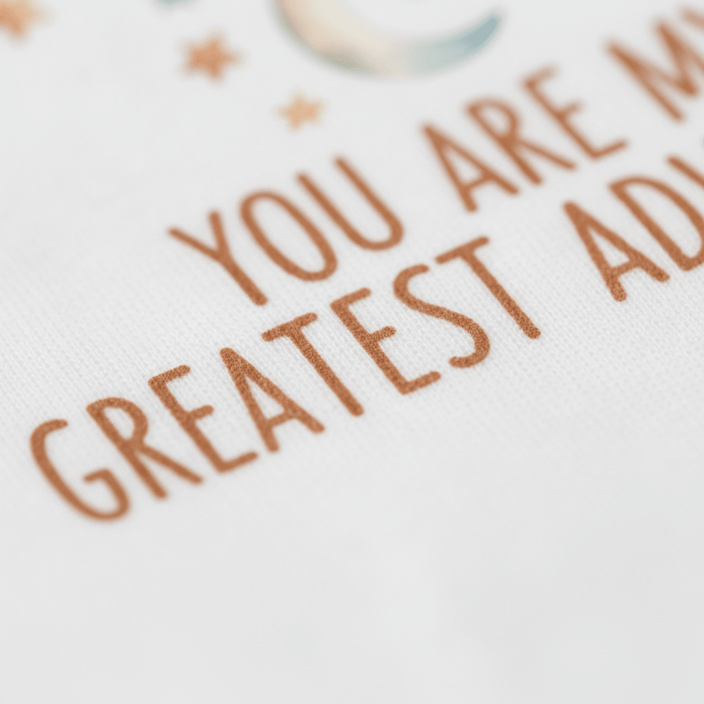 Close-up of brown text on white fabric, partially reading YOU ARE MY GREATEST ADVI..., with small stars and a crescent moon above the inscription. The Toodleroos “You Are My Greatest Adventure Baby Onesie – Hot Air Balloon” captures a sense of wonder and love in every detail, making it the perfect adventure-themed gift for welcoming a new baby.