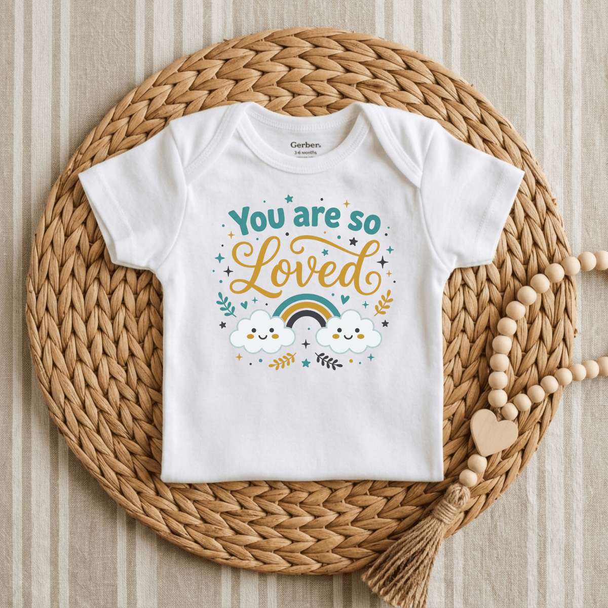 The Toodleroos “You Are So Loved Onesie – Cute Baby Rainbow Bodysuit” features the heartfelt message “You are so loved” alongside clouds, stars, and a rainbow. Displayed on a woven mat next to a wooden bead garland with a heart, this charming onesie is perfect for anyone wanting cute baby onesies and makes an ideal gender-neutral baby gift.