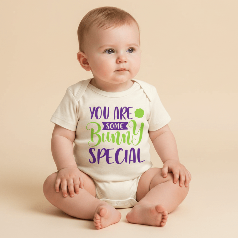 A baby sits on a beige backdrop, dressed in the Toodleroos “You Are Some Bunny Special Baby Onesie – Easter Bodysuit,” which features the phrase “You are some bunny special” in playful purple and green letters. Gazing calmly to the right, the baby highlights how this unisex bodysuit from Toodleroos makes a delightful spring baby shower gift.