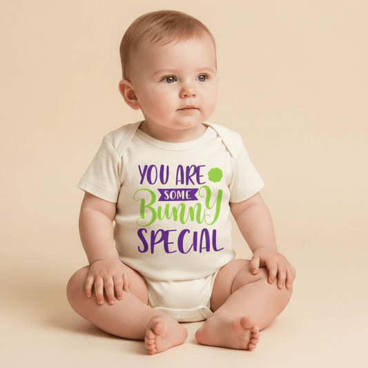 A baby sits on a beige backdrop, dressed in the Toodleroos “You Are Some Bunny Special Baby Onesie – Easter Bodysuit,” which features the phrase “You are some bunny special” in playful purple and green letters. Gazing calmly to the right, the baby highlights how this unisex bodysuit from Toodleroos makes a delightful spring baby shower gift.