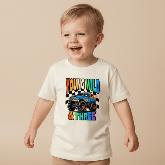 A smiling young boy wears the Toodleroos Young Wild & Three Toddler Shirt – Monster Truck 3rd Birthday Tee, featuring a cartoon monster truck and colorful “Young Wild & Three” text—the perfect shirt for any toddler who loves monster trucks on their 3rd birthday.