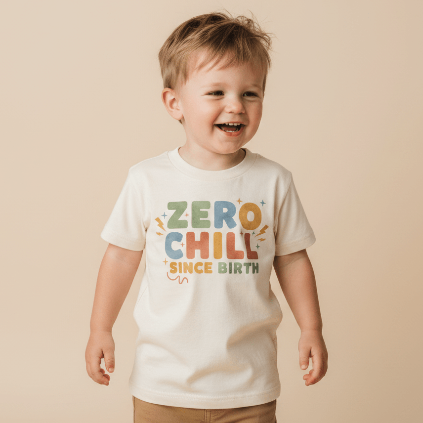 A smiling young boy wears the Toodleroos Zero Chill Since Birth Toddler Shirt – a funny retro tee – featuring colorful text, while standing against a beige background.